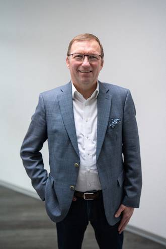 Teammitglied mit Brille in grauem Sakko und weißem Hemd, selbstbewusste Haltung mit Hand in der Hosentasche vor neutralem Hintergrund im Business-Portrait.