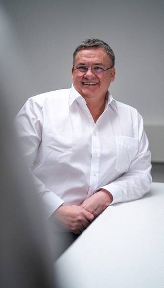 Teammitglied mit grauen Haaren in weißem Hemd, freundlich lächelnd mit verschränkten Armen vor hellem Hintergrund im professionellen Portrait.