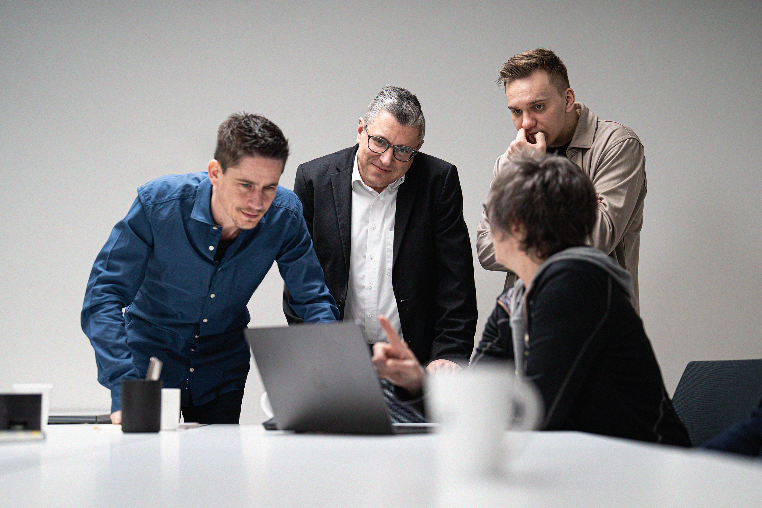 Vier Teammitglieder um einen Laptop versammelt, Person in blauem Hemd steht und erläutert etwas am Bildschirm, während zwei Kollegen in dunkler Business-Kleidung aufmerksam zuschauen, Farbaufnahme an weißem Konferenztisch.