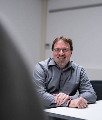 Teammitglied mit Brille und Bart in grauem Pullover, freundlich lächelnd an einem Bürotisch sitzend in heller Büroumgebung im Portrait.