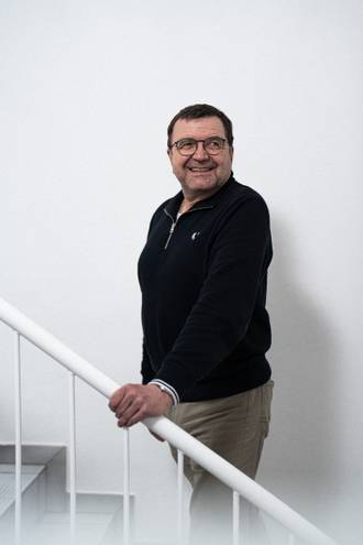 Teammitglied mit Brille in schwarzem Pullover und beiger Hose, lächelnd und mit der Hand am Treppengeländer vor weißer Wand stehend.