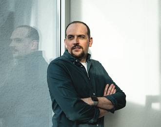 Teammitglied mit kurzen Haaren und Bart in schwarzem Oberteil mit verschränkten Armen, selbstbewusste Pose vor hellem Hintergrund im Portrait.