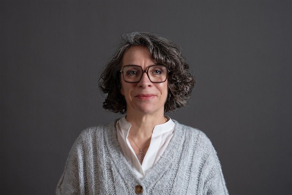 Teammitglied mit lockigen schulterlangen Haaren und Brille in weißer Bluse unter hellgrauer Strickjacke, freundlich lächelnd vor dunkelgrauem Hintergrund in professionellem Farbportrait.