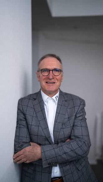 Business Software Spezialist mit Brille in grauem Sakko und weißem Hemd, freundlich lächelnd in professioneller Pose vor hellem Büro-Hintergrund.