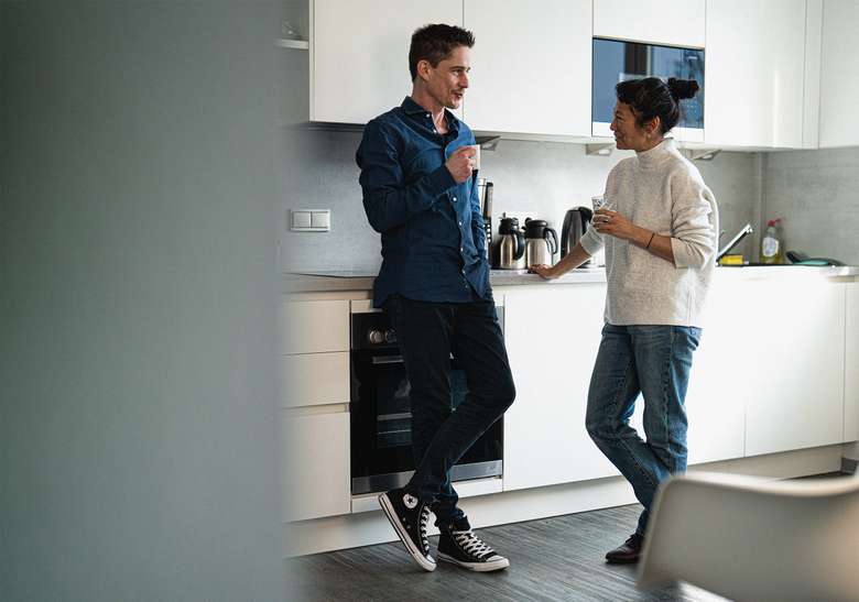 Zwei Teammitglieder in einer modernen Büroküche, Person in dunkelblauem Hemd öffnet einen Küchenschrank, während eine Person in hellem Oberteil vorbeigeht, Farbaufnahme in minimalistischem Küchenambiente.