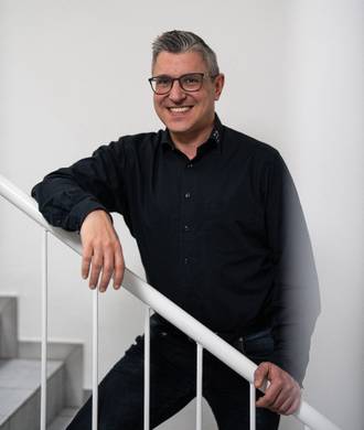 Teammitglied mit Brille und grauem Haar in schwarzem Hemd, entspannt mit einer Hand am weißen Treppengeländer lehnend vor hellem Hintergrund.