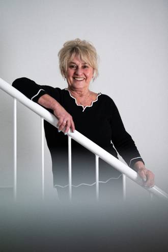 Teammitglied mit Brille und blonden kurzen Haaren in schwarzem Oberteil, freundlich lächelnd an einem weißen Treppengeländer stehend im Business-Portrait.