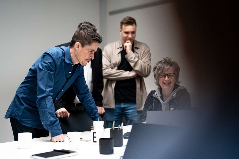 Business-Meeting mit vier Teammitgliedern an einem Konferenztisch, wobei eine Person im blauen Hemd stehend Unterlagen präsentiert, während die anderen aufmerksam zuhören und Notizen machen.