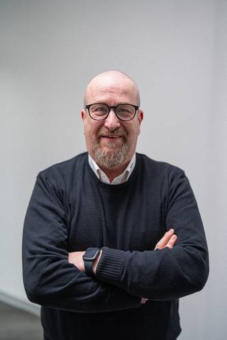IT-Experte mit Brille und Vollbart in schwarzem Pullover und weißem Hemd mit verschränkten Armen, selbstbewusste Haltung im Business-Portrait.