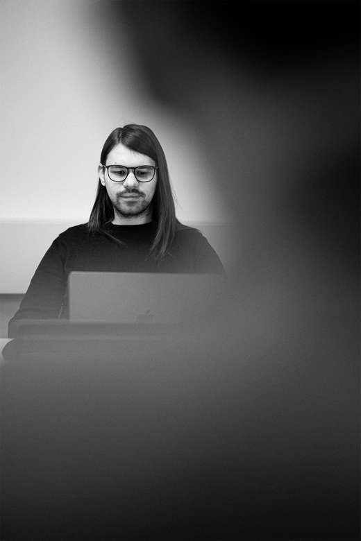 Teammitglied mit langen dunklen Haaren in schwarzem Oberteil konzentriert an einem Computer arbeitend,  Schwarz-Weiß-Portrait mit dramatischer Beleuchtung.