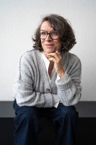 Teammitglied mit Brille und schulterlangen Haaren in grauer Strickjacke und dunklem Rock, nachdenkliche Pose mit Hand am Kinn im Schwarz-Weiß-Portrait.