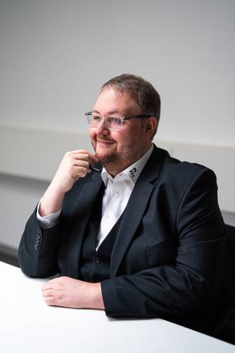 Teammitglied mit Brille in dunklem Sakko und weißem Hemd, nachdenklich mit Hand am Kinn an einem Bürotisch sitzend im Business-Portrait.