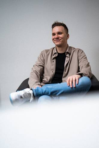 Teammitglied mit kurzen Haaren in beiger Jacke und Jeans, lächelnd in entspannter Sitzposition mit Dokumentenrolle auf weißem Hintergrund.