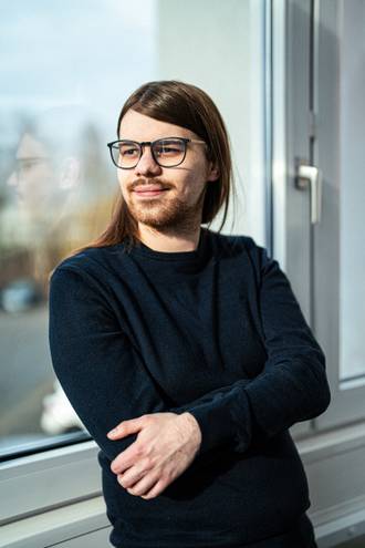 Teammitglied mit Brille und schulterlangen Haaren in schwarzem Pullover, lächelnd mit verschränkten Händen vor hellem Fensterbereich im Business-Portrait.