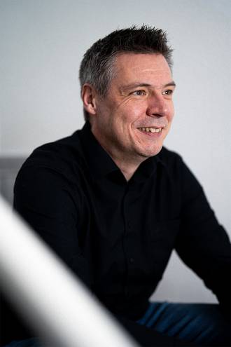 Teammitglied in schwarzem Hemd mit kurzen Haaren, freundlich lächelnd im Seitenprofil vor hellem Hintergrund im professionellen Portrait.