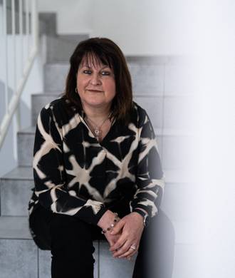 Teammitglied mit schulterlangen dunklen Haaren in schwarz-weißem Muster-Pullover und schwarzer Hose, freundlich lächelnd auf einer weißen Treppe sitzend.