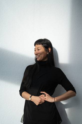 Teammitglied mit schulterlangen Haaren in schwarzem Rollkragenpullover, nachdenkliche Pose mit verschränkten Händen vor hellem Hintergrund im Portrait.