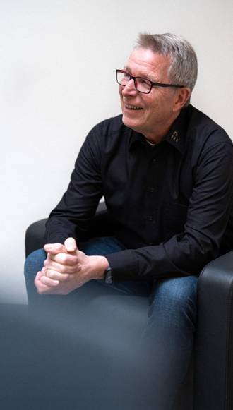 Teammitglied mit Brille und grauen Haaren in schwarzem Hemd, entspannt sitzend mit gefalteten Händen vor weißem Hintergrund im Business-Portrait.