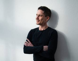 Business Software Berater in schwarzem Pullover mit verschränkten Armen vor hellem Hintergrund in professioneller Seitenansicht.