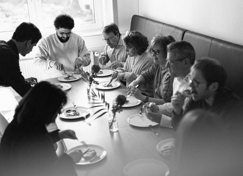 Team bei einer gemeinsamen Mahlzeit am Konferenztisch, mehrere Personen sitzen zusammen mit Tellern und Essen vor sich, informelle Besprechungsatmosphäre, Schwarz-Weiß-Aufnahme von oben.