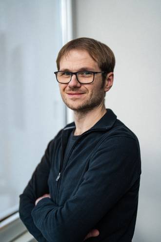 Teammitglied mit Brille und kurzen Haaren in dunkelblauem Pullover mit verschränkten Armen, freundlich lächelnd vor hellem Fensterbereich im Business-Portrait