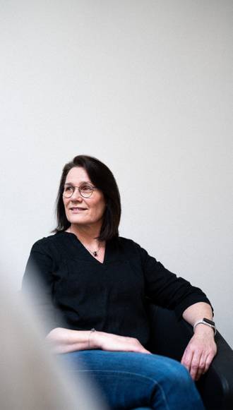 Teammitglied mit Brille und dunklen Haaren in schwarzem Oberteil, seitlich auf weißem Stuhl sitzend mit entspannter Körperhaltung vor hellem Hintergrund.