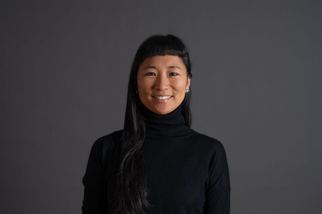 Teammitglied mit langen dunklen Haaren in schwarzem Rollkragenpullover, freundlich lächelnd vor dunkelgrauem Hintergrund in professionellem Farbportrait.